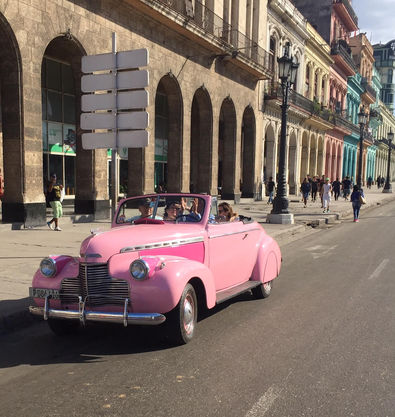 The pink convertible is iconic in Havana because of its color and luxury car status. Note the colorful buildings in the background.
 
 